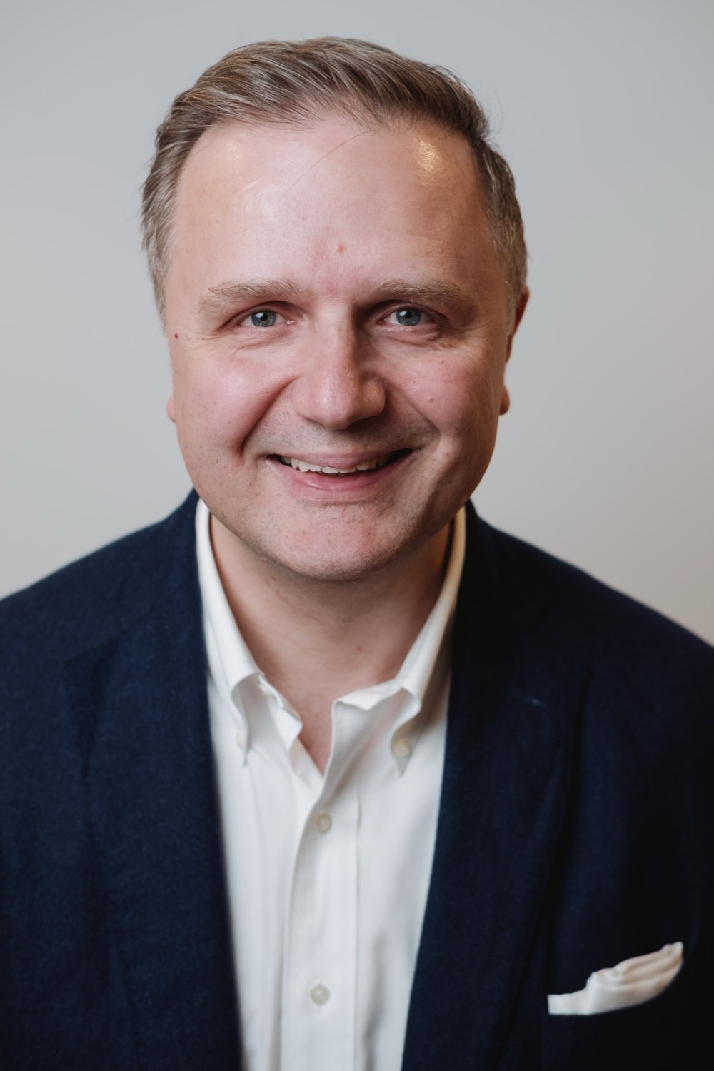 Professional headshot of a smiling man in a navy blazer on a pale backdrop